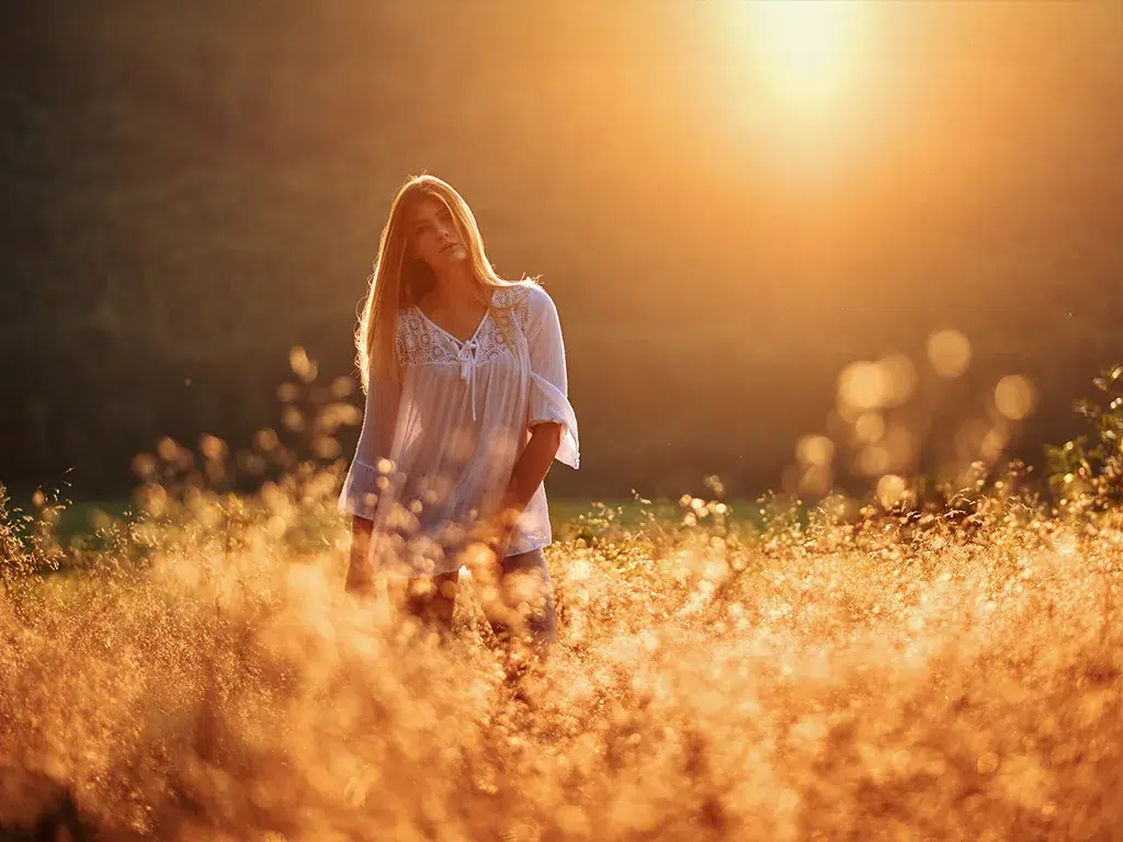 Frau in weißer Bluse steht in goldenem Sonnenuntergang inmitten blühender Wiese mit Lichteffekten